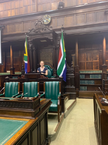 A woman sits in a large chambers seat with the flag of South Africa on either side of her